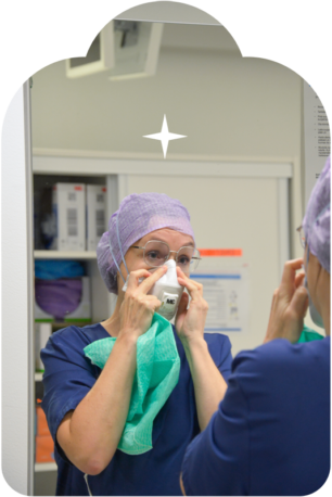 Sairaanhoitaja katsoo peiliin ja asettaa maskia kasvoilleen, päässään hiusverkko ja kädessä vihreä suoja-asu