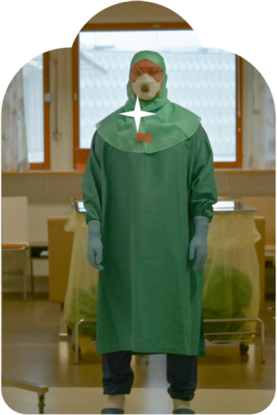 Henkilö seisoo teho-osastolla vihreässä suoja-asussa, maski kasvoillaan. Taustalla muovilla suojattuja sairaalalaitteita